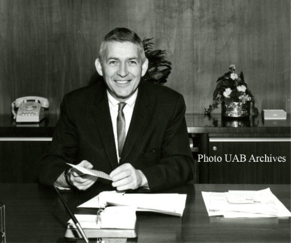 A black and white image of an older white man in a suit (Dr. Volker) smiling for a photo at his desk in his office wit hthe words to the right,